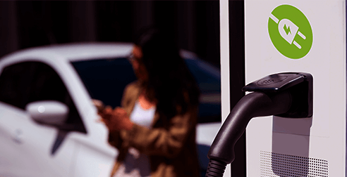 Tomada carregando veículo híbrido e ao fundo mulher branca olhando o celular encostada em veículo branco