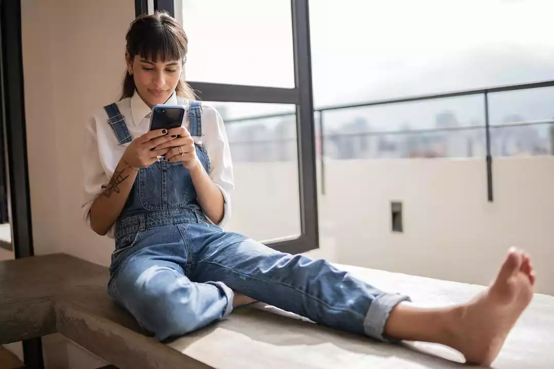 Foto de uma mulher mexendo no celular, sentada em uma varanda. Ela está vestindo um macacão jeans e camisa branca. Ela tem cabelo preto, franja e tatuagem no braço.